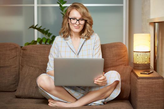 A young woman in glasses works remotely on a laptop in her apartment. A cute girl during a video conference with her colleagues at home. A female student with a smile listening to an online lecture.