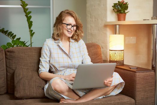 A woman in a dress sitting on the couch with legs crossed works remotely on a laptop in her apartment. A girl with braces watching a webinar. A female student in glasses listening to an online lecture