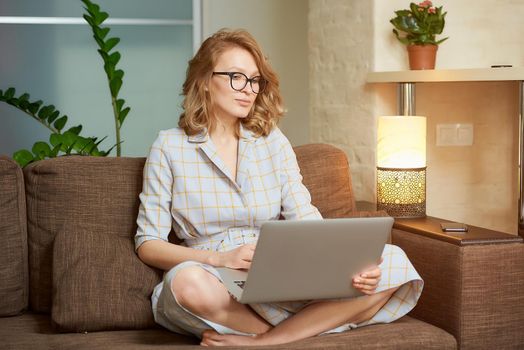 A woman in a dress sitting on the couch with legs crossed works remotely on a laptop. A pretty girl watching a webinar in her apartment. A female student in glasses listening to an online lecture.