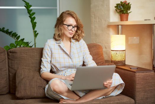 A woman in a dress sitting on the couch with legs crossed works remotely on a laptop. A serious girl with braces watching a webinar. A female student in glasses listening to an online lecture.