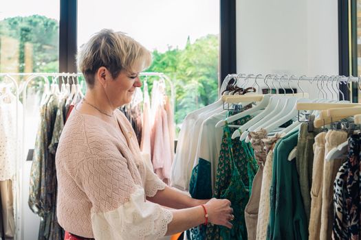 blonde mature woman happy and enjoying a day of shopping, choosing a pink dress from a rack, shopping in a fashion shop clothes. shopping concept. natural light from the shop window, sun rays, display with clothes, rack, shop window, clothes, Horizontal.