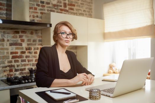 A young woman in glasses works remotely on a laptop in her kitchen. A blond girl listening to her colleagues on a video conference at home. A lady studying a lesson online on a webinar.