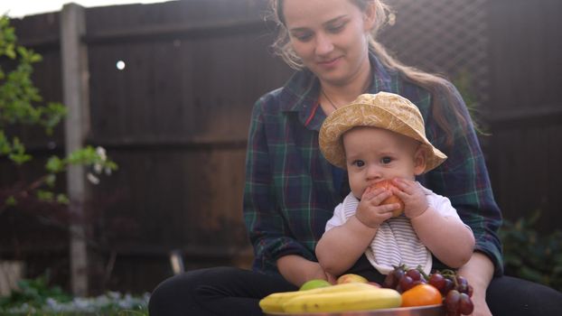 Happy Young Cheerful Mother Holding Baby Eating Fruits On Green Grass. Mom Adorable Infant Child Playing Outdoors With Love In Backyard Garden. Little Kid With Parents. Family, Nature, Ecology Concept.