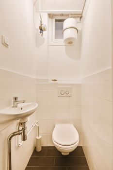 Modern toilet and ceramic sink installed on white tiled walls under small window and boiler in narrow restroom at home