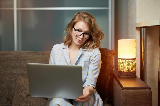 A young woman in glasses works remotely on a laptop in her apartment. A lady during a video conference at home. A pretty female student listening to an online lecture.