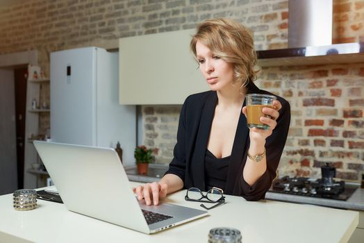 A young woman works remotely on a laptop in her kitchen. A girl calmly browsing news on the internet on her computer at home. A lady holds a glass of coffee preparing for a lecture on a video call..