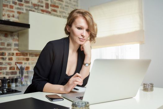 A young woman works remotely on a laptop in a kitchen. A beautiful girl calmly listening to a report of a colleague at a video conference at home. A female teacher preparing for an online lecture