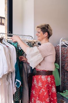 business woman, fashion shop owner, hanging clothes on hangers in her business. shopping concept. leisure concept. natural light from the shop window, sun rays, display with clothes, clothes rack, shop window, clothes, vertical.