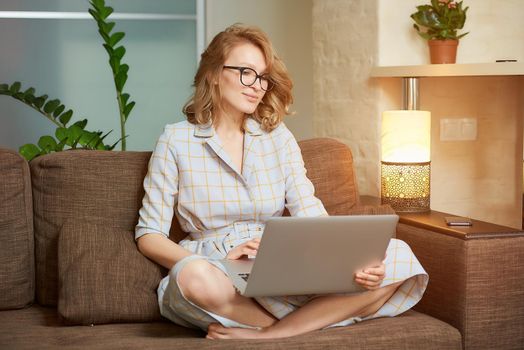 A woman in a dress sitting on the couch with legs crossed works remotely on a laptop. A happy girl watching a webinar in her apartment. A female student in glasses listening to an online lecture.