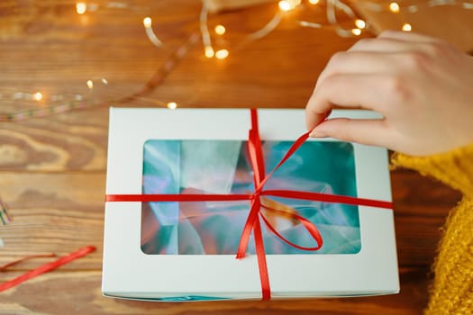 First-person view of woman's hand in knitted sweater opening Christmas surprise. Girl pulls ribbon on gift box. Wooden table with garland. New Year's holiday mood.