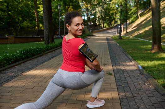 View from the back to an African woman doing lunges while exercising outdoor, smiles cutely with beautiful toothy smile looking at camera, enjoying her active and healthy lifestyle