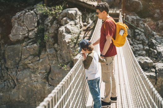 Father and son looking afar from the rope bridge in the mountains when hiking. Casually dressed tourists child school boy and his dad with yellow backpack crossing the canyon through rope bridge.