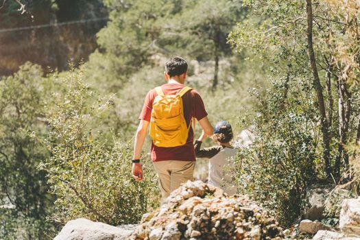 Rear view of tourists school boy and his dad walking a stone footpath in spring forest. Child boy and father wearing casual clothes and yellow backpack while hiking in summer greenwood leaf forest.