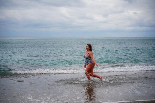 A plump woman in a bathing suit enters the water during the surf. Alone on the beach, Gray sky in the clouds, swimming in winter