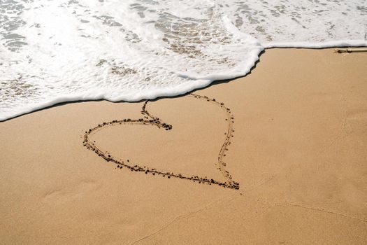 Boy in yellow rubber boots drawing heart shape on sand at the beach. School kid touching water at autumn winter sea. Child having fun with waves at the shore. Spring Holiday vacation concept.