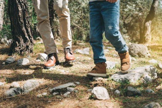 Close-up view of tourists school boy and his dad feet boots walking a stone footpath in spring forest. Child boy and father wearing hiking boots while walking in summer greenwood forest.