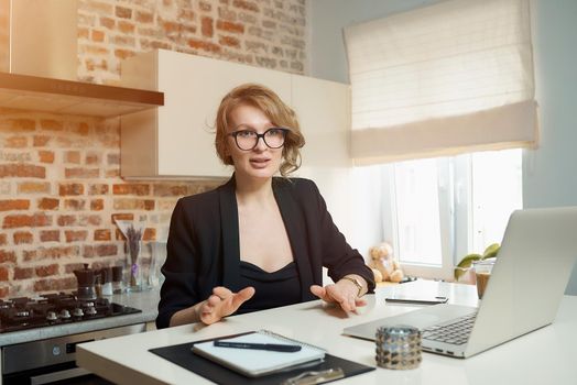 A young woman in glasses works remotely on a laptop in her kitchen. A blond girl gesticulating discusses with her colleagues on a video conference at home. Lady teaching a lecture online on a webinar