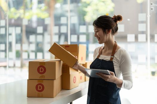 Young business woman working online e-commerce shopping at her shop. Young woman seller prepare parcel box of product for deliver to customer. Online selling, sme, e-commerce