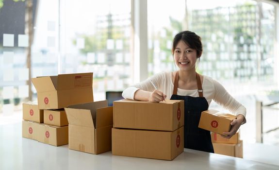 Young Asian small business owner working at home office, taking note on purchase orders. Online marketing packaging delivery, startup SME entrepreneur or freelance woman concept.