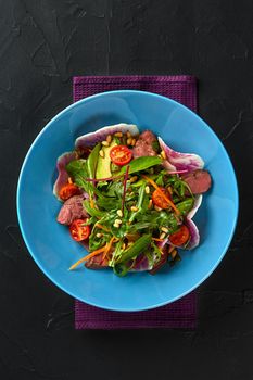 Spicy beef slices meat salad with tomatoes, parsley, radish and salad leaves spinach on black texture background table. Still life