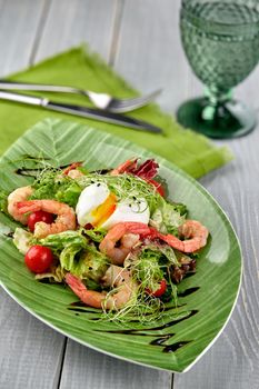 Salad from shrimp with lettuce, arugula, eggs, tomatoes, olive oil and balsamic vinegar on a green plate on a wooden table