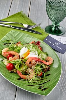 Salad from shrimp with lettuce, arugula, eggs, tomatoes, olive oil and balsamic vinegar on a green plate on a wooden table