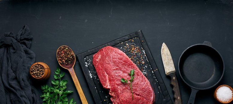 raw beef tenderloin lies on a wooden cutting board and spices for cooking on a black table, top view