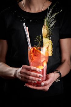 Pink fruit cocktail or lemonade in the hands of a woman in a black T-shirt. Pineapple with orange.