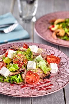 Healthy mixed Greek salad served on a pink plate with silver fork containing crisp leafy greens, microgreen, feta, onion, tomato and sliced beef. Selective focus. Lunch. Still life