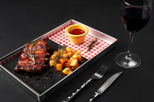 Delicious barbecued ribs seasoned with a sauce, potatoes and served with herbs on a metal tray, on black background. Serving a ready meal in a restaurant, cafe, bar, pub. Still life. Copy space