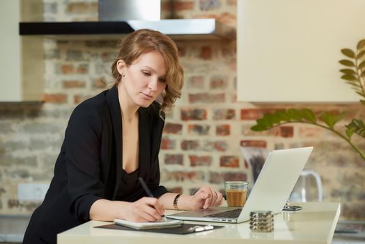 A young woman works remotely in her kitchen. A charming girl doing notes in the notebook during a video conference at home. A female teacher preparing for an online lecture.