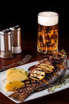 Grilled carp fish with rosemary potatoes, lemon and beer on a wooden table. Black background. Selective focus