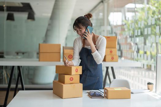 Young Asian small business owner working at home office, taking note on purchase orders. Online marketing packaging delivery, startup SME entrepreneur or freelance woman concept.