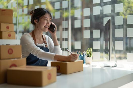 Young Asian small business owner working at home office, taking note on purchase orders. Online marketing packaging delivery, startup SME entrepreneur or freelance woman concept.