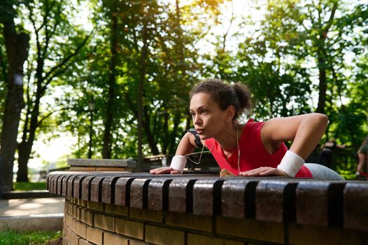 Strong confident beautiful African American fitness girl in athletic sportswear doing push up, performing full body functional workout in the urban park