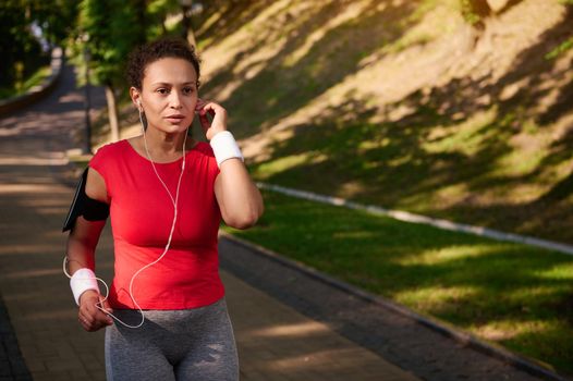 Middle aged African American determined sportswoman, runner listens to music on earphones while running along the city park treadmill. Jog, cardio workout, healthy and active lifestyle concept