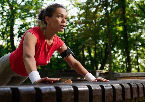 Close-up of a determined Hispanic female athlete, muscular build woman in bright sportswear performing push ups from wooden bench in the city park during functional cross training in the morning