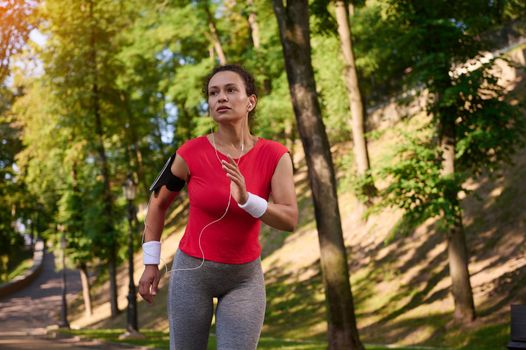 Attractive strength muscular multi ethnic woman with healthy beautiful aesthetic fit body in tight sportswear with earphones and smartphone holder running on the footpath in the city park. Sport