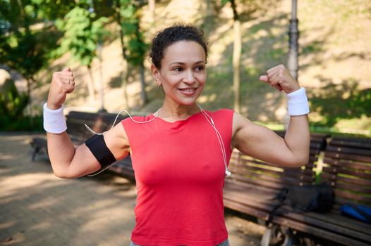 Charming confident active African American woman, athlete, sportswoman clenching fists, tensing arm and biceps muscles, smiling standing on the city park during sports training. Go beyond your limits.