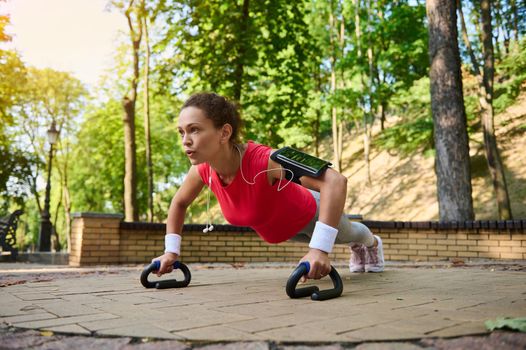 Go beyond your limits. Athletic middle-aged active woman in bright sportswear and smartphone holder doing push ups during functional full body workout in urban landscape park. Keep your body fit