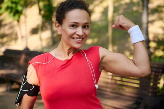Go beyond your limits. Active multi ethnic middle aged athlete muscular sportswoman smiling beautiful toothy smile and showing her toned biceps to camera during workout. Sport and healthy body concept