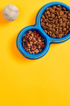 Dry pet food in bowl with a ball on yellow background top view. Still life. Copy space. Flat lay