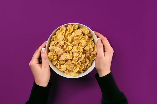 Female hands holding bowl with healthy breakfast, closeup. Bowl with cornflakes on the colorful background. Purple background, top view. Copy space. Still life. Flat lay