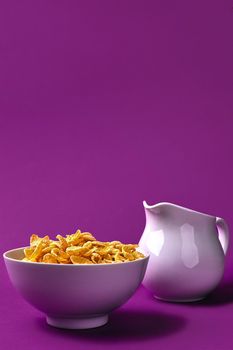 Bowl with corn flakes, jug of milk on purple background. oncept of a healthy breakfast. Copy space. Still life. Flat lay