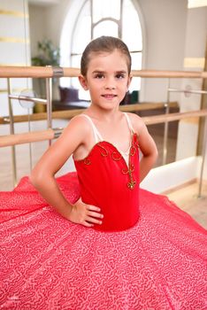 Cute portrait of a beautiful little ballerina in a performance red dress with pink tutu. She smiles because she is happy to become a professional ballerina in the future