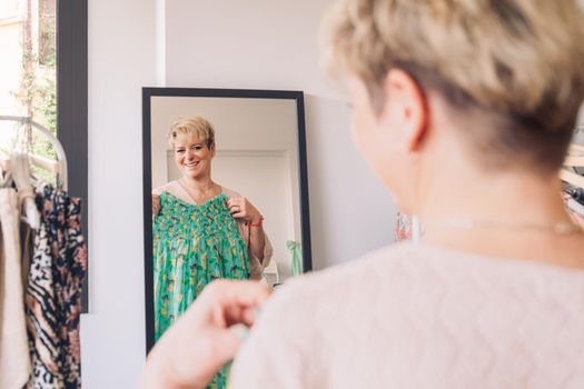 mature woman testing a dress in front of a mirror. blonde female shopping in a fashion shop. shopping concept. leisure concept. Natural light, sunbeams, display, clothes rack, clothes, horizontal view, space to copy.