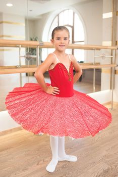 Cute portrait of a beautiful little ballerina in a performance red dress with pink tutu. She smiles because she is happy to become a professional ballerina in the future