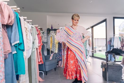 mature woman testing a dress in front of a mirror. blonde female shopping in a fashion shop. shopping concept. leisure concept. Natural light, sunbeams, display, clothes rack, clothes, vertical view, space to copy.