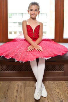 Full length portrait of a beautiful little ballerina in a performance red dress sitting by the window and dreaming to become professional ballet dancer