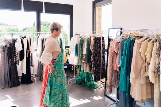 mature woman testing a dress in front of a mirror. blonde female shopping in a fashion shop. shopping concept. leisure concept. Natural light, sunbeams, display, clothes rack, clothes, horizontal view, space to copy.